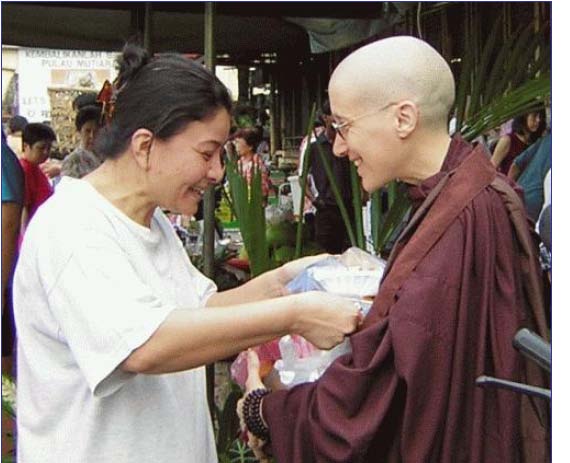 [A smiling woman places a packet of food in Ayya Medhanandi's bowl]